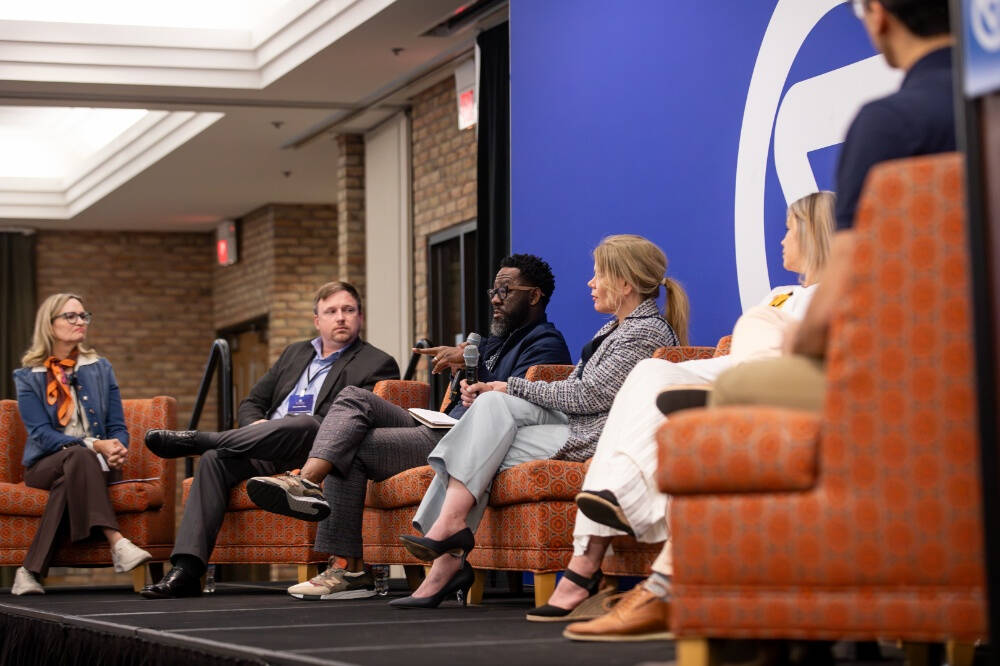 A group of people, both men and women are seated on orange patterned chairs on a stage, engaged in a panel discussion. The background features a large blue wall with a white logo, and the setting appears to be a conference or professional event.